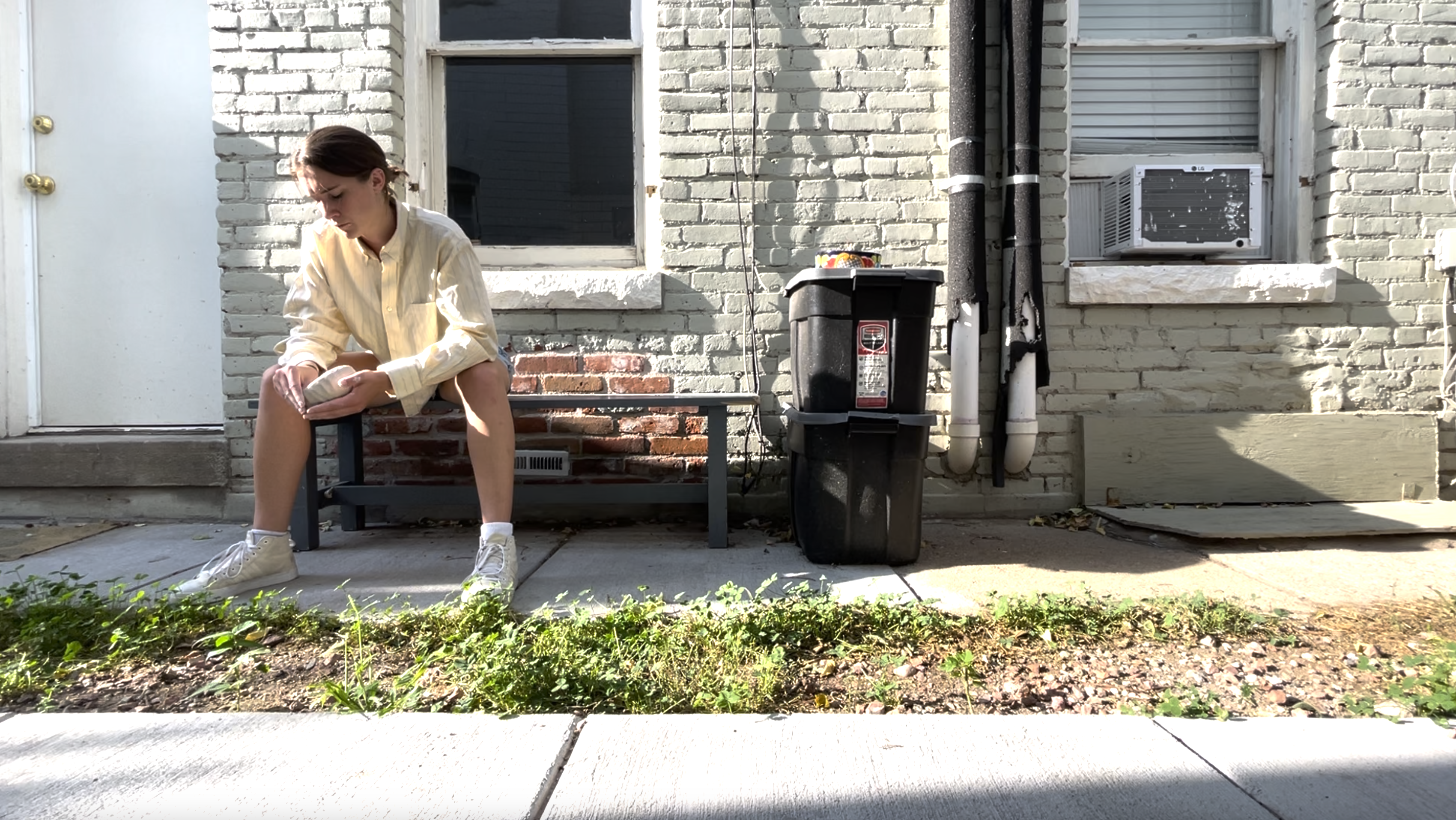 Sarah sits opn a bench outside of a white brick building making a pinch pot.